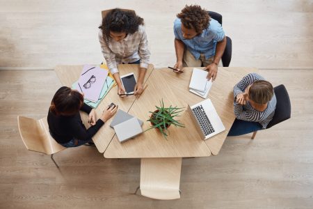 View from above of group young professional entrepreneurs sitting at table in coworking space, discussing profits of last team project, using laptop, digital tablet and smartphone